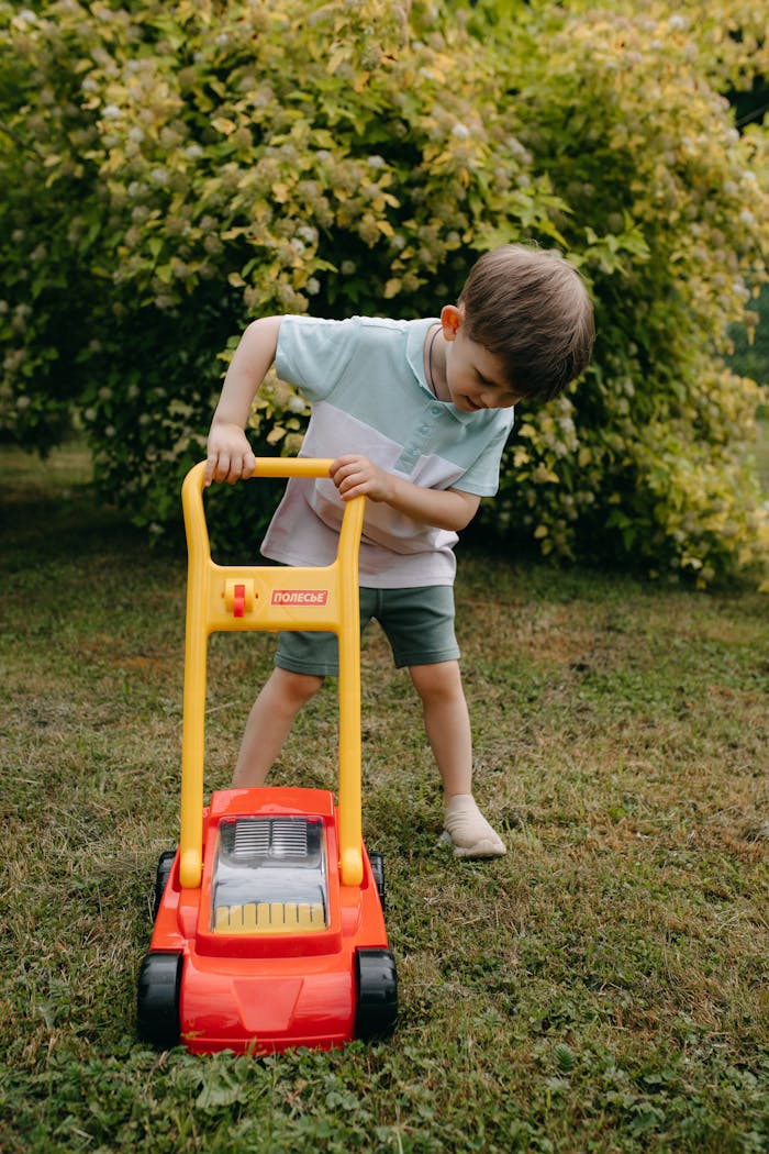Young boy enjoys playing with a red toy lawn mower in a green garden setting on a sunny day.