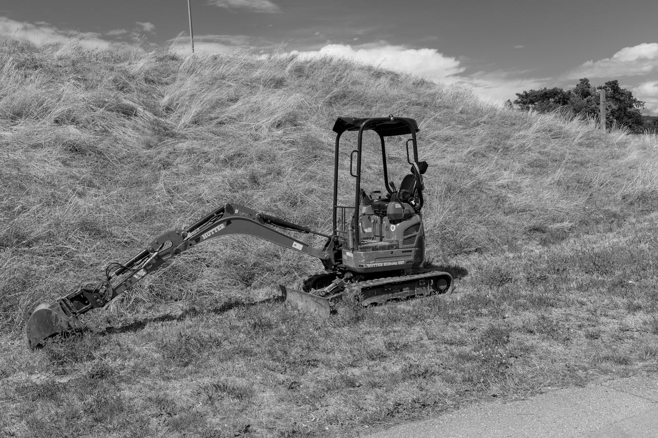 A black and white image of a mini excavator on a grassy hill. Industrial and serene contrast.